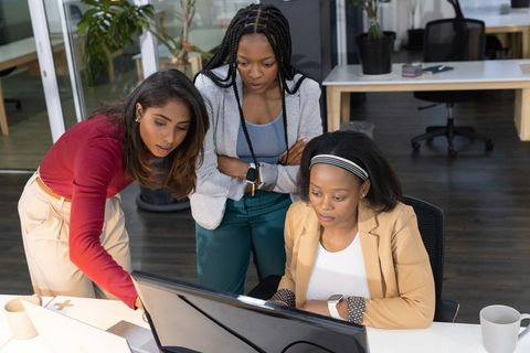 Three diverse women collaborating and pointing at desktop monitor in modern open office
