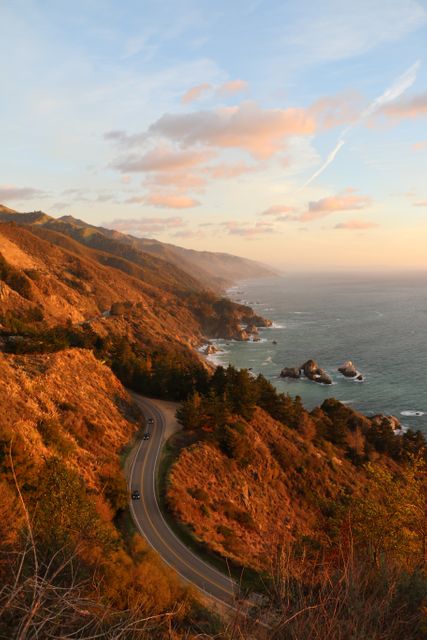 This beautiful photograph depicts a winding coastal highway during sunset, showcasing a stunning ocean view with mountains and cliffs in the background. The warm evening light enhances the dramatic scenery, making it perfect for travel blogs, adventure magazines, nature documentaries, romantic getaway promotions, and road trip advertisements.