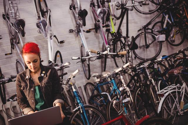 Bicycle mechanic with red hair using laptop in workshop filled with bicycles. Ideal for content related to bicycle repair, maintenance services, modern technology in traditional trades, and small business operations.