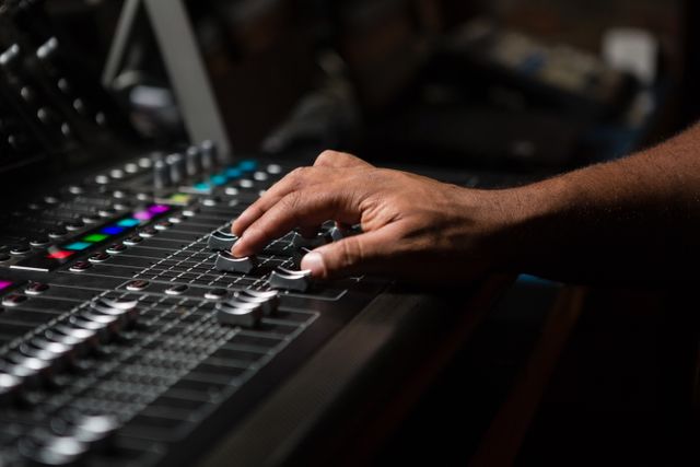Hand of a male audio engineer adjusting controls on a sound mixer in a recording studio. Ideal for use in articles or advertisements related to music production, sound engineering, professional audio equipment, and the music industry.