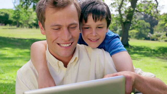 Father and son enjoying leisure time in park, sharing an ebook on a tablet. Perfect for themes related to family bonding, technology interaction, outdoor activities, and quality leisure time.