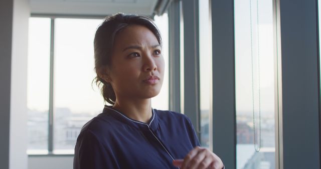 Asian woman in business attire standing by window in office. Ideal for use in business, corporate, and professional development contexts. Can be used in presentations, blogs about career growth, and workplace environment features.