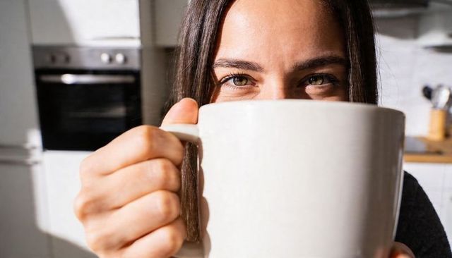 Woman holding oversized white mug close to face while enjoying morning sun in kitchen
