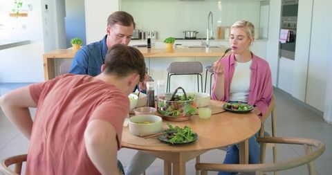 Family sharing fresh salad around round wooden dining table in bright contemporary kitchen