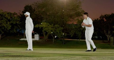 Diverse male cricketers preparing bowling delivery under floodlights at dusk on grass pitch