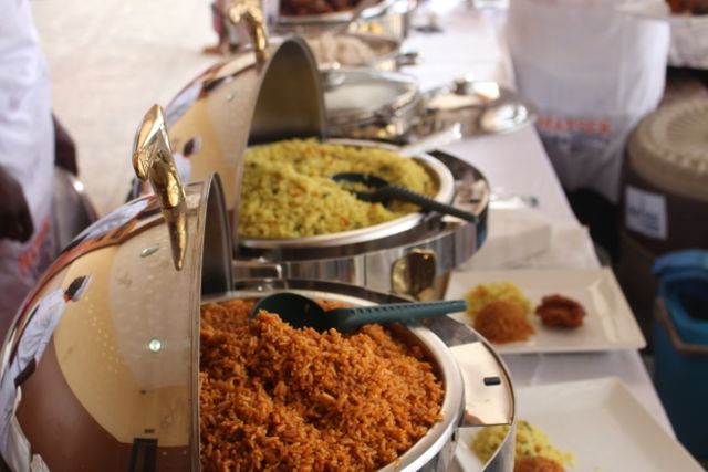 This image portrays a buffet setup featuring various traditional African rice dishes at a catered event. The food trays are filled with vibrant, appetizing options, capturing the essence of African culinary culture. Ideal for use in materials related to catering services, event planning advertisements, or cultural celebration promotions, this visual showcases the richness and appeal of African cuisine.