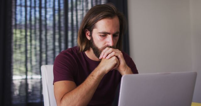 Young man with a beard deeply focused on work, facing laptop screen, likely dealing with important tasks. Useful for depicting concentration, home office setup, remote work environment, or digital work scenarios.