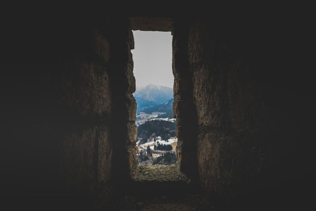 A view of a mountain landscape seen through the window of a rocky cave frame. This serene and scenic shot emphasizes the contrast between the dark cave interior and the bright, snowy mountain peaks outside. Suitable for travel blogs, adventure websites, nature magazines, and environmental awareness campaigns.