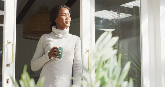 Young woman standing by the doorway, holding a coffee mug and enjoying the morning sunlight. She is dressed casually in a cozy sweater, radiating a sense of peace and relaxation. Ideal for use in lifestyle blogs, wellness articles, advertisements for clothing brands, or promoting coffee and relaxation. Could also be used for articles or visual content related to morning routines and healthy living.