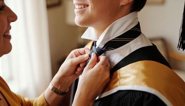 Parent fastening graduate's tie while preparing cap and gown with gold academic hood