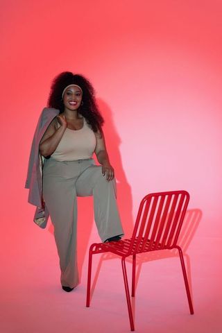 Confident african american woman posing with blazer and red chair against pink backdrop