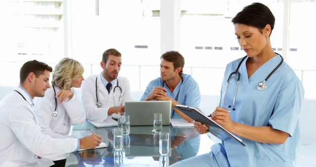 Healthcare team engaging in a clinical discussion around a table with a doctor reviewing notes on a clipboard. Ideal for illustrating collaborative efforts in patient care, teamwork in healthcare settings, medical staff meetings, and healthcare planning.