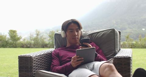 Asian man relaxing on wicker lounge chair in backyard using phone and tablet
