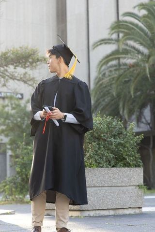 Asian graduate standing on campus wearing black gown and yellow tassel holding diploma