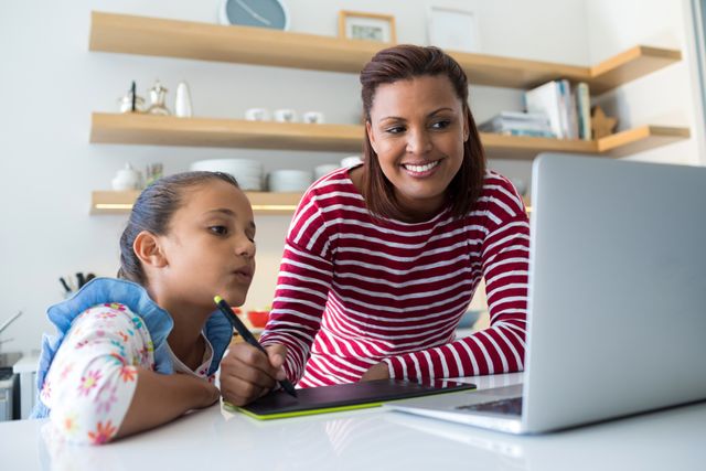 Mother and daughter using graphic tablet while looking at laptop in kitchen