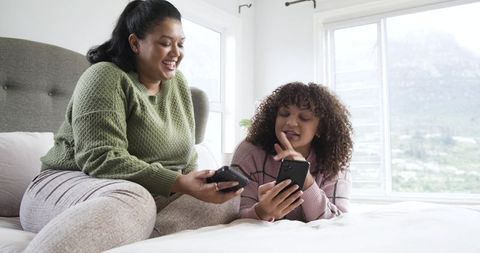 Two women sharing smartphones and laughing on bed in bright modern bedroom interior