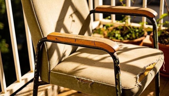 Sunlit vintage armchair with frayed seat and wooden arms on balcony with potted plants
