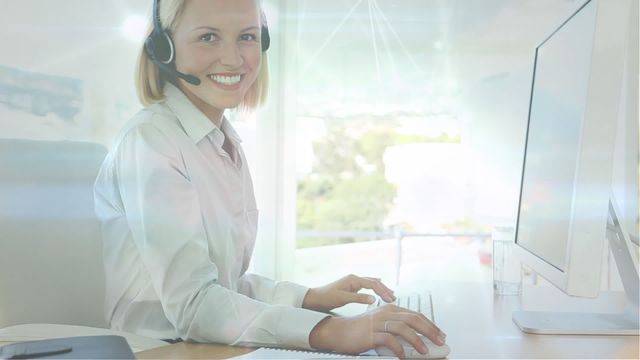 A woman in a professional setting wearing a headset smiles while working on a desktop computer with a digital networking overlay. This videory illustrates modern customer service and communication technology, making it suitable for use in presentations, websites, and marketing materials related to call centers, tech support, and professional customer service environments.