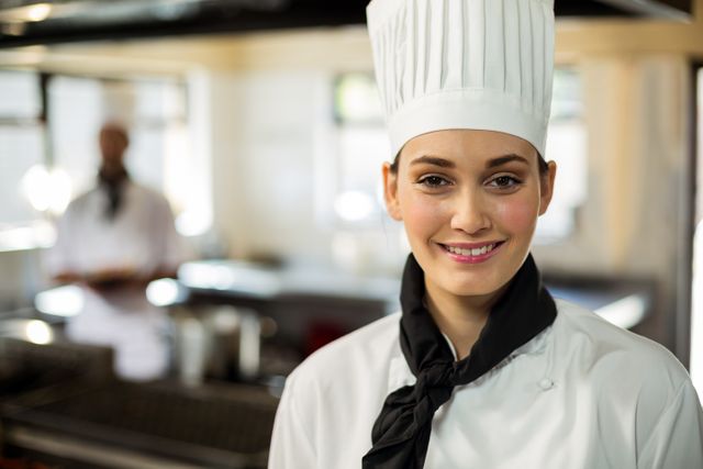 This image depicts a smiling female chef standing in a commercial kitchen, wearing a traditional chef uniform and hat. Ideal for use in culinary blogs, restaurant websites, hospitality industry promotions, cooking school advertisements, and food-related publications. The background shows a professional kitchen environment, adding authenticity to the setting.