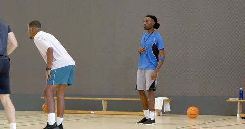 Basketball coach supervising drills on indoor court with players, bench and balls