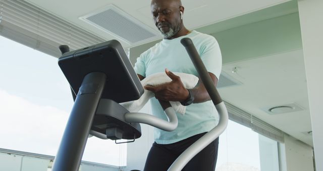 Man working out on elliptical machine attentively holding towel. Useful for fitness promotion, healthy living advertisements, senior fitness programs. Showcases dedication to staying healthy, suitable for gym interiors, exercise and wellness content.