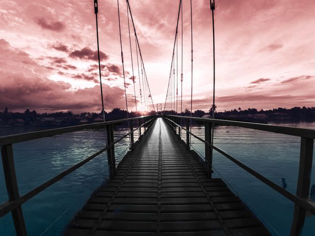 Suspension bridge extends into sunset horizon creating dramatic and peaceful scenery. Wooden walkway above tranquil ocean waters with silhouettes of distant palm trees. Ideal for travel, adventure, tropics or inspiring journey concepts.