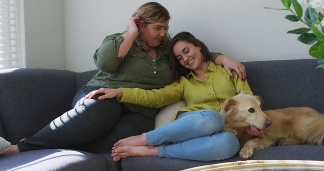 Two women are sitting on a couch at home, showing affection for each other while their golden retriever dog is resting beside them. They are dressed casually and appear relaxed and happy, creating a warm and inviting family scene. Perfect for use in topics related to family, companionship, LGBTQ+ representation, or home lifestyle.