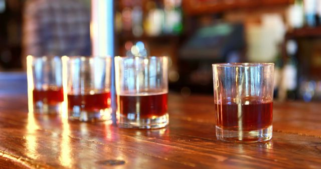 Four glasses with amber liquid on weathered wooden bar counter, bright bar ambiance in background, displaying socializing, celebration, or party. Perfect for pubs, bars, drinking posters, or entertainment promotions.