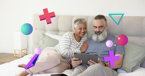 Mature couple relaxing on bed sharing tablet, smiling, cozy home technology moment