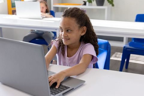 Smiling elementary girl typing on laptop in bright classroom, digital learning focus