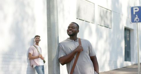 Two friends leaning on urban pole smiling with casual backpacks on sunlit sidewalk