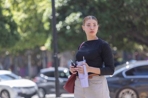 African american woman standing on sidewalk holding lavender cup and phone checking traffic in city