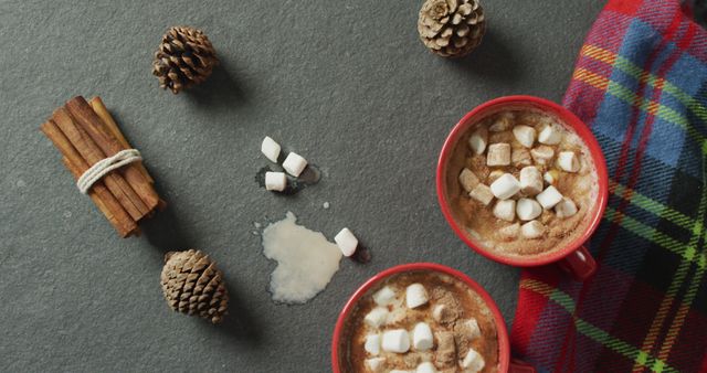 Warm hot chocolate with marshmallows on dark stone surface. Surrounded by cinnamon sticks, pine cones, with spilled drink and plaid blanket for cozy winter vibe. Perfect choice for promoting winter drinks, festive menus, holiday-themed blogs, or cozy home settings.