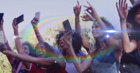 Friends raising smartphones at outdoor festival with rainbow light flare and joyful expressions