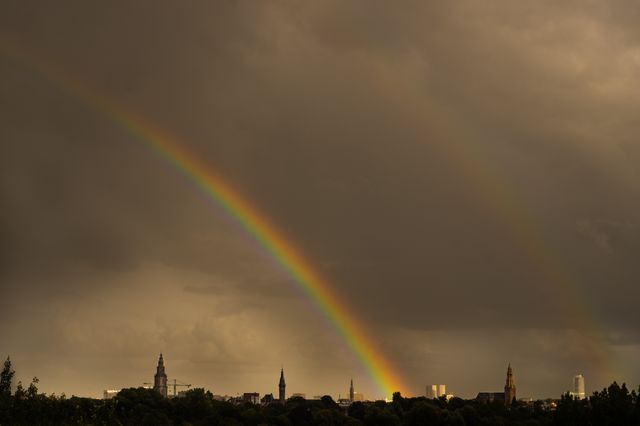 Vibrant rainbow arching over a city skyline during dusk. Dark storm clouds provide a dramatic backdrop, enhancing the colors of the rainbow and adding contrast to the scene. Visible structures and spires showcase architectural elements. Suitable for themes related to weather, natural phenomena, urban landscapes, and atmospheric beauty.