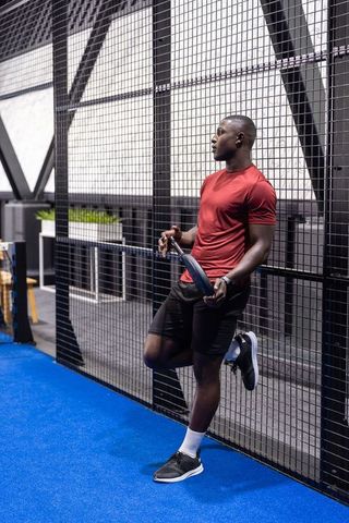 Urban athlete leaning against mesh fence holding tennis racket on blue indoor gym floor