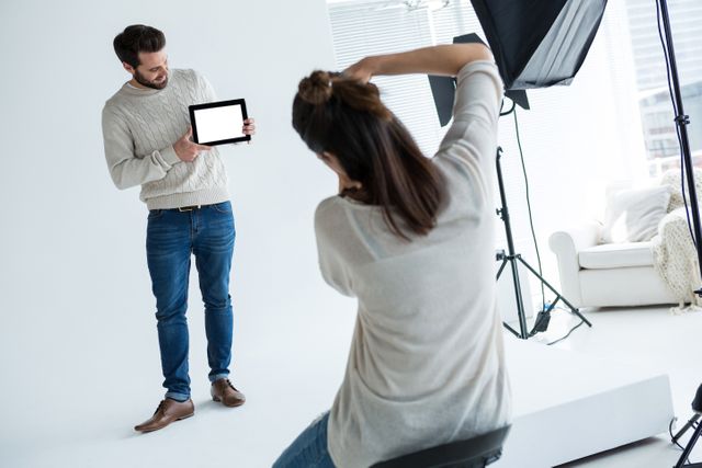 Male model holding a tablet and posing in a professional photography studio. Photographer capturing the scene with studio lighting and equipment. Ideal for use in articles about fashion, technology, professional photography, and modeling.