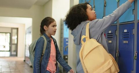 Teen girls opening lockers in school hallway carrying backpacks and wearing casual clothes