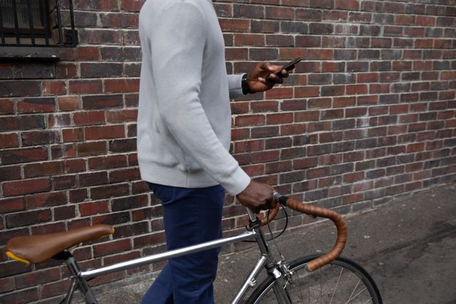 Mid section of smart casually dressed African American man out and about in the city streets during the day wheeling a silver racing bicycle with drop handlebars and using smartphone