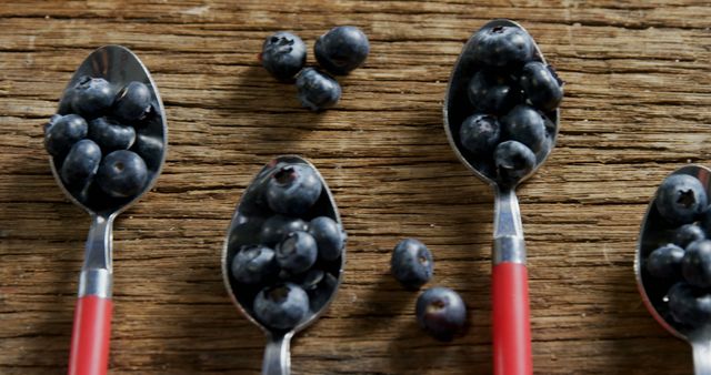 Spoons with red handles are filled with plump blueberries on a rustic wooden surface, with some berries scattered around. The image captures a simple yet artistic presentation of fresh fruit, evoking a sense of wholesome and natural eating.