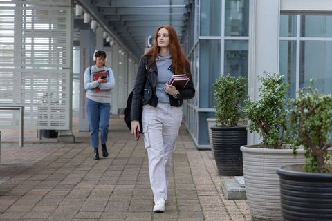 Redhead student walking with notebooks along modern campus walkway, carrying backpack