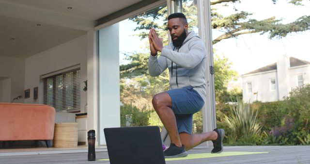 A young man is practicing yoga at home with the help of an online class on his laptop. He is wearing a hoodie and shorts, kneeling on a yoga mat positioned in front of the laptop. The room looks modern and spacious with natural light streaming in, and the view outside shows a lush, green garden. This image can be used to promote online fitness classes, healthy lifestyle habits, home workouts, and mental well-being through yoga.