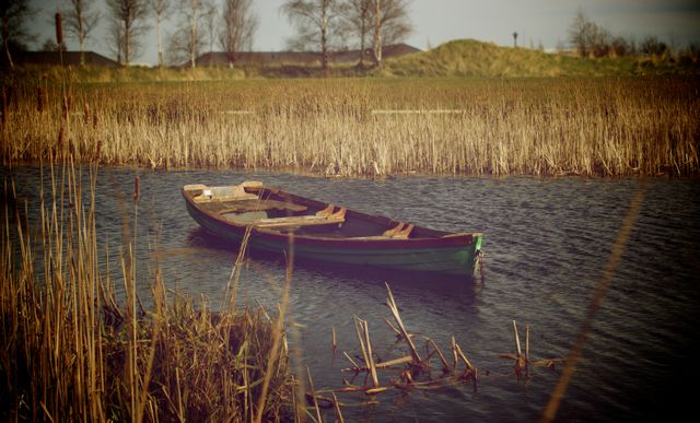 Depicting an abandoned wooden boat floating on a calm lake surrounded by reeds, this image captures a rustic and serene countryside scene. It evokes feelings of solitude and tranquility, ideal for use in nature-themed projects, travel blogs, environmental campaigns, or artistic portfolios conveying peace and quietness.