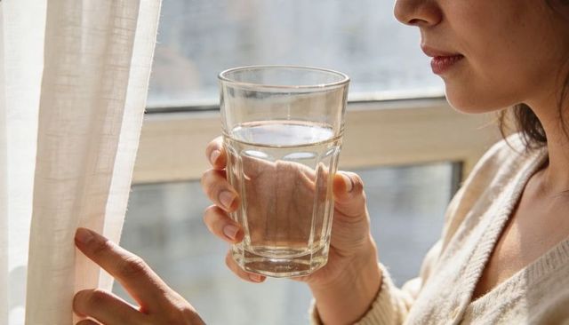 Sunlit morning woman holding glass of water pulling curtain in V-neck sweater