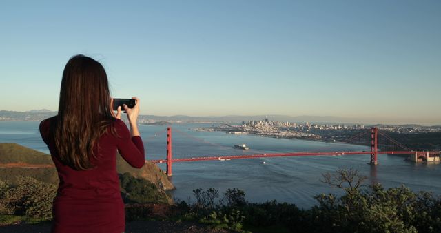 Rear view of a young Caucasian woman with long dark hair walking in the Golden Gate National Recreation Area and stopping to take pictures of the Golden Gate Bridge and the San Francisco Bay Area with a smartphone 