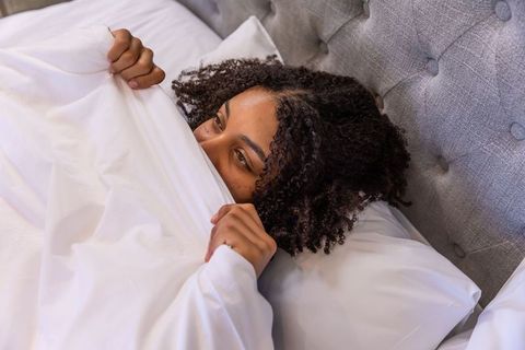 African american woman snuggling under duvet on tufted gray headboard, cozy bedroom