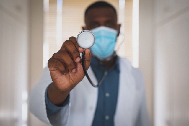 African american male doctor wearing face mask holding stethoscope to camera. medical professional at work during coronavirus covid 19 pandemic.