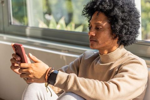 Sunlit african american man relaxing by window using smartphone in beige sweater