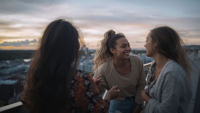 Three friends laughing on rooftop terrace at sunset overlooking city skyline