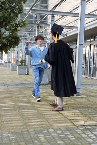 Campus reunion: graduate in cap and gown greeting friend on modern university walkway